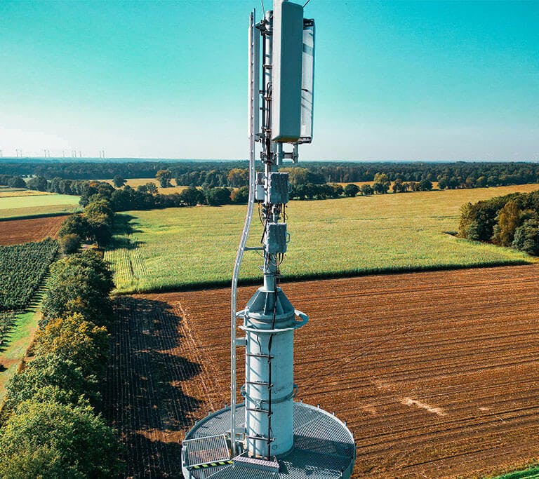 A cell tower can be seen in the foreground of a vast, agricultural landscape on a sunny day.