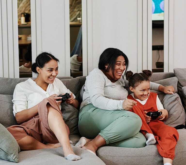 A family of three sits on a couch, playing a video game together while holding controllers and laughing.