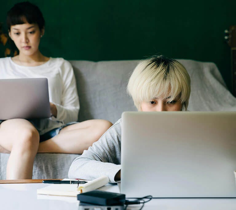 One person sits on a couch and uses a laptop near another person who is sitting on the floor and also using a laptop that’s on a table next to a charging smartphone.