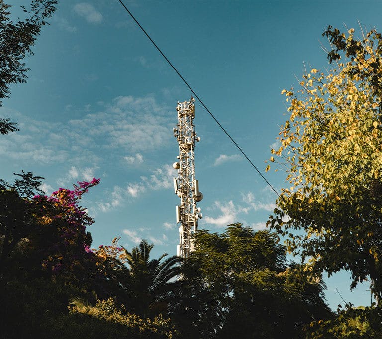 A cell tower emits a signal against a blue sky with beautiful trees in the foreground.