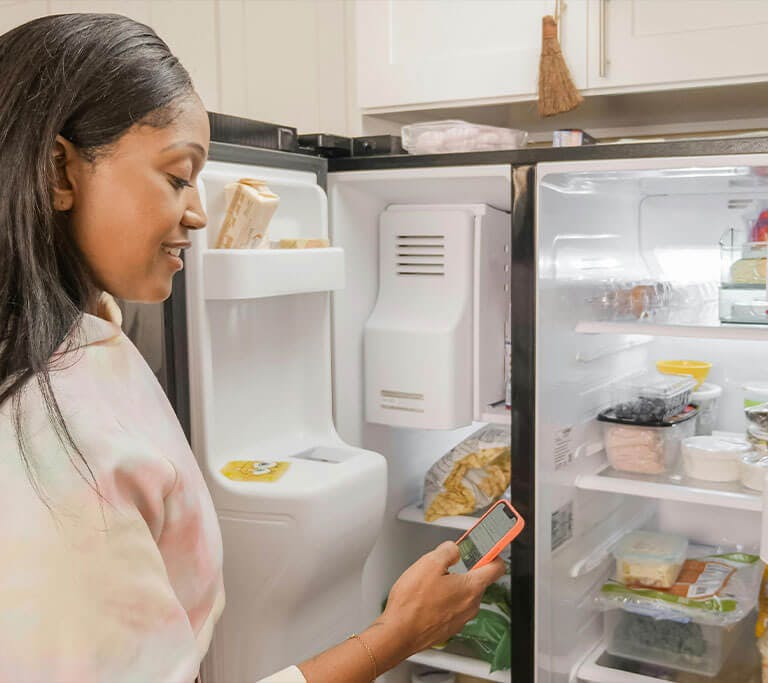 A woman holding a cell phone in front of an open refrigerator