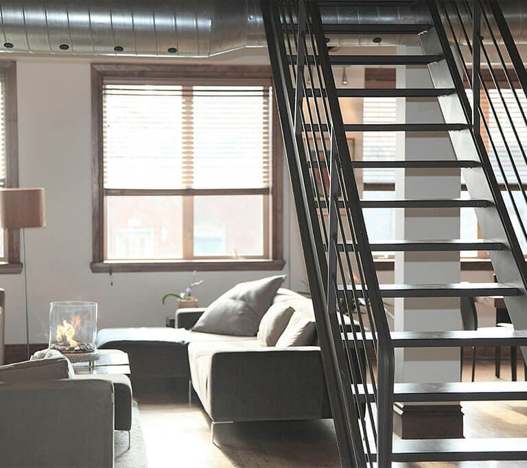 A modern loft interior features a stylish living room with a sleek staircase, cozy furniture, a warm fireplace, and large windows adorned with elegant blinds.