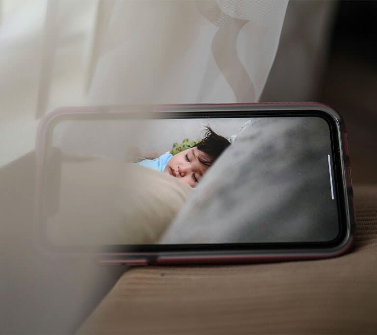 A smartphone displays the view from a baby monitor in the next room and shows a sleeping baby with brown hair.