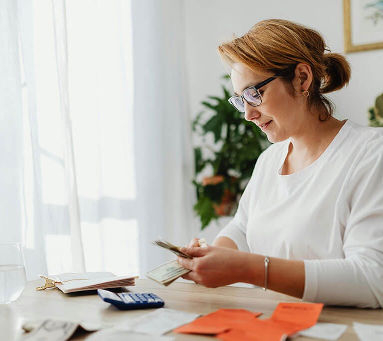 A person in a white shirt and glasses smiles in her home office as she counts dollar bills.