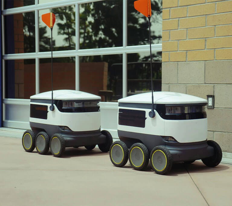 A pair of white and black, six-wheeled delivery robots with large antennas move past a brick university building.