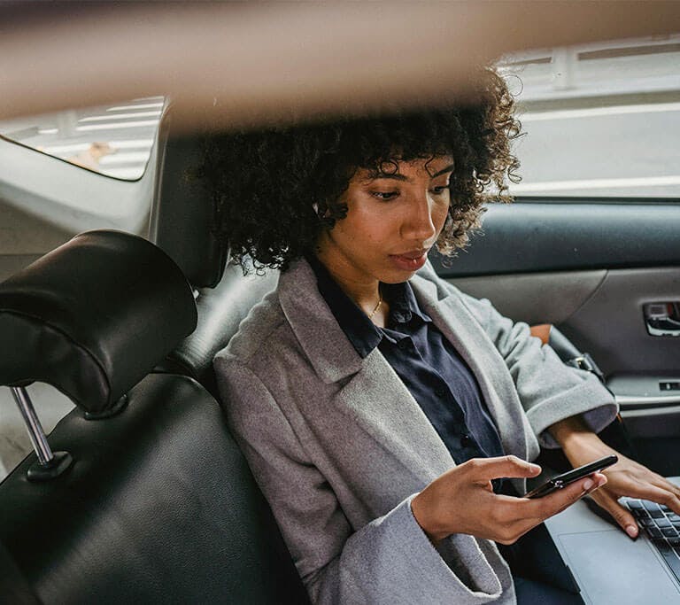 woman wearing a grey blazer is holding her phone and using a laptop in the backseat of a car with black, leather seats.