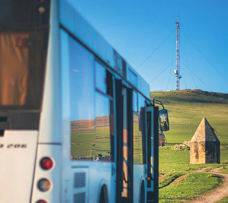 A cell tower can be seen in the background as a white bus drives over rolling green hills under a bright, blue sky in a rural area.