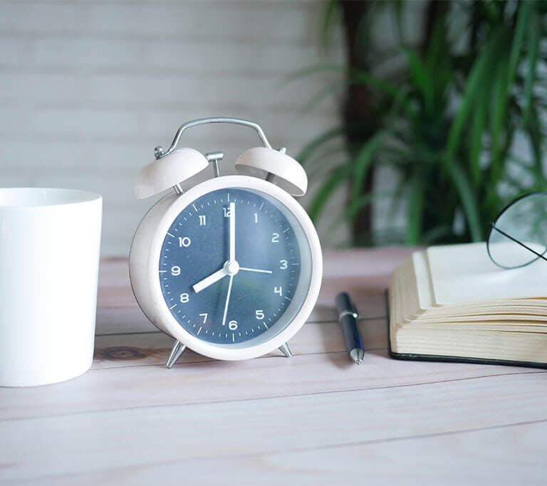 An alarm clock with bells on it sits on a desk next to a book, glasses  and a container.