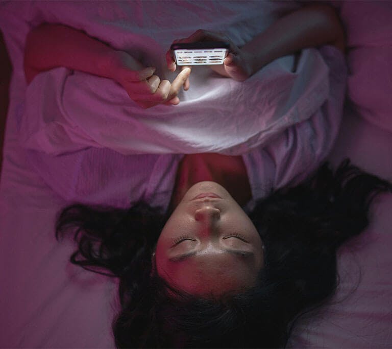 A person lies in bed under white sheets while changing settings on their phone.