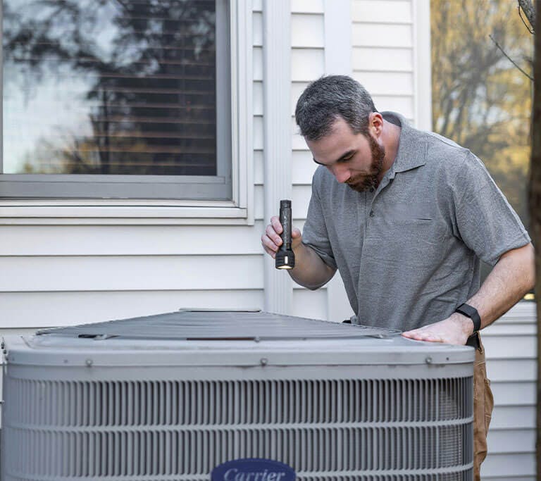 A person wearing a grey polo is pointing a flashlight at an HVAC unit outside of a home.