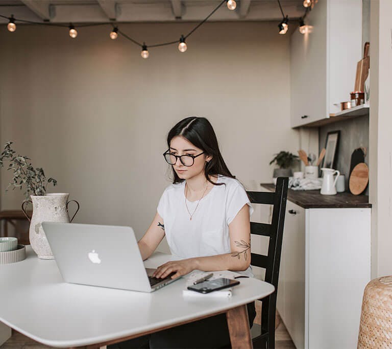A person wearing glasses sits at a white table and types on a silver MacBook in a minimalist room.