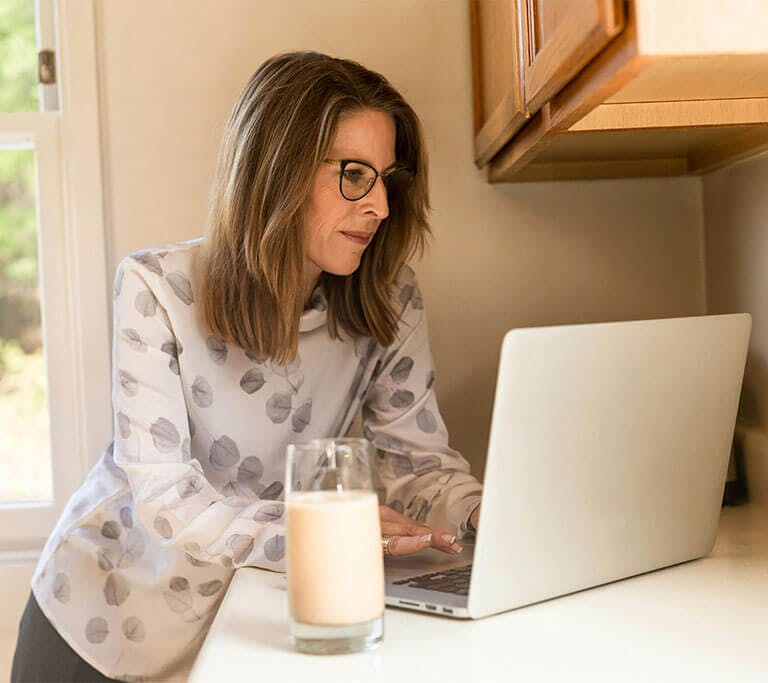 A person wearing a white shirt with a grey leaf print pattern uses their laptop in their kitchen.