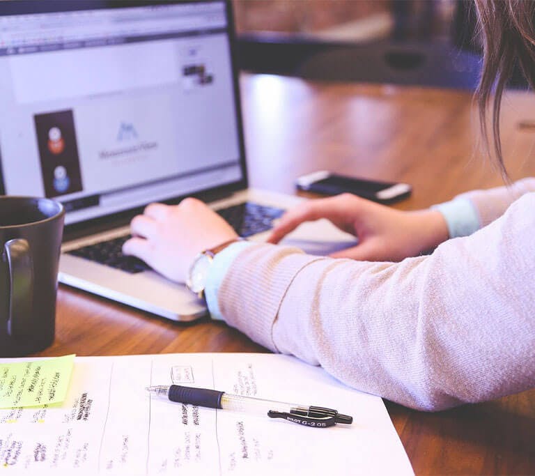 Woman typing on a laptop at a wooden table. She has a paper and pen beside her as well as a coffee cup.