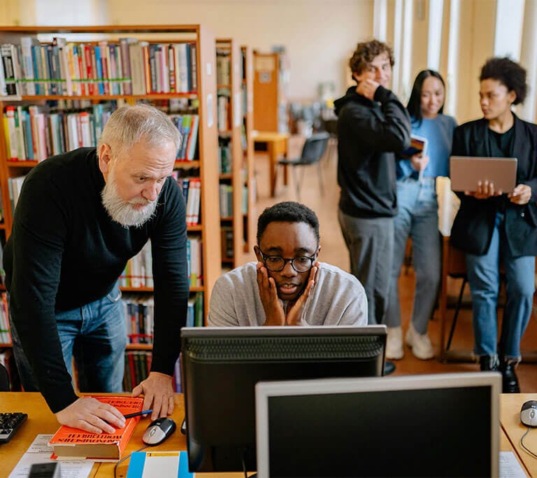 An older person with a white beard leans over to assist a younger person, who appears to be distressed and is working at a desktop computer in a library. In the background, a group of three people talk and use laptops and tablets near tall bookshelves.