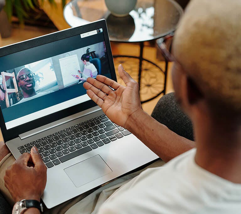 A person sits with a laptop on their lap during a video call, gesturing toward the screen where two other callers appear in separate windows.