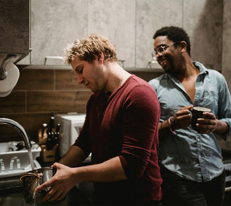 Two people are standing in a modern kitchen, while one fills a silver pot with hot water and the other holds a mug, smiling.