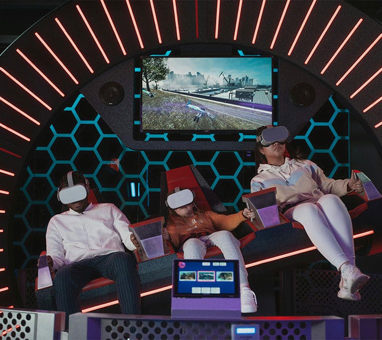 A family of three sit in a futuristic-looking booth with VR headsets on as they play a VR game.