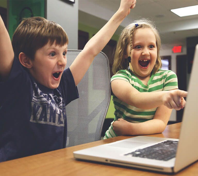 Two children, who look extremely excited, are sitting at a wooden table in a room with green and white walls in front of a silver laptop. One child points at the screen and the other has their arms raised in a victory pose.