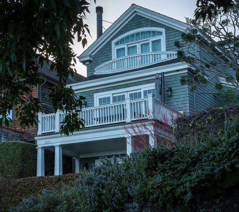 A three-story grey house with two balconies and lots of windows surrounded by vegetation.