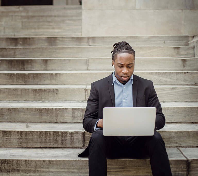 A businessperson wearing a suit sits on steps and appears frustrated while using a silver laptop.
