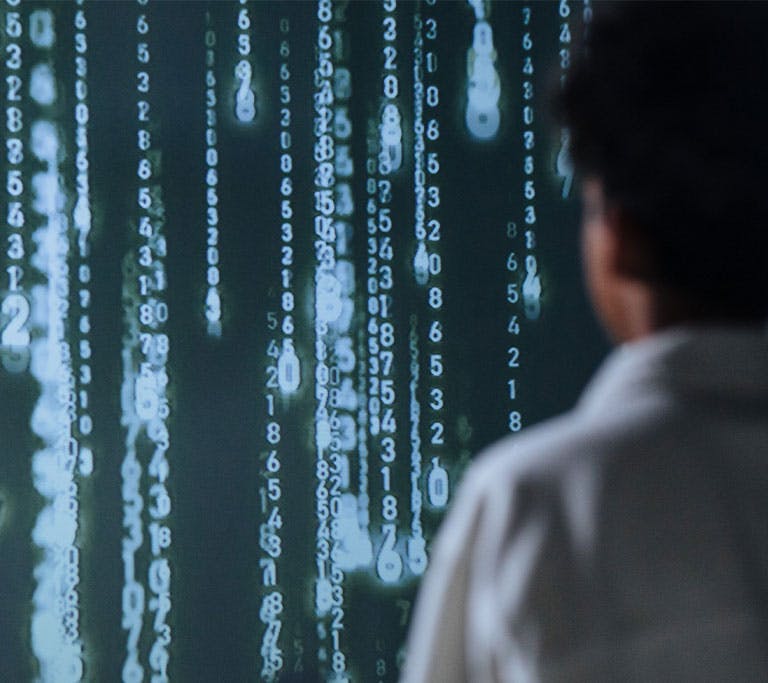 Photograph showing a person in a white shirt facing a digital screen filled with cascading vertical streams of glowing numbers and symbols in blue and white hues. The scene evokes themes of data analysis, coding, or cybersecurity, highlighting the interac