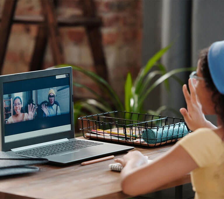 A child sitting at a wooden desk near a houseplant lifts a hand to wave at two people who are also waving in a video call on a laptop screen.
