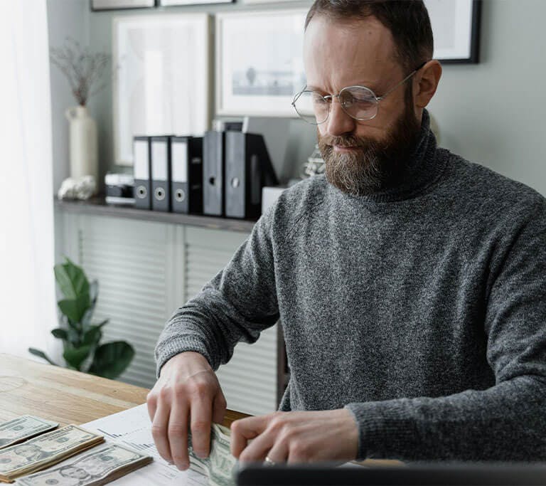 A person wearing circular glasses and a grey sweater puts cash into different piles on a light-brown wooden table.
