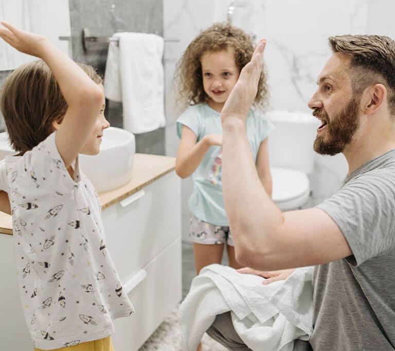 A parent high-fives two young children inside a bright, clean bathroom.