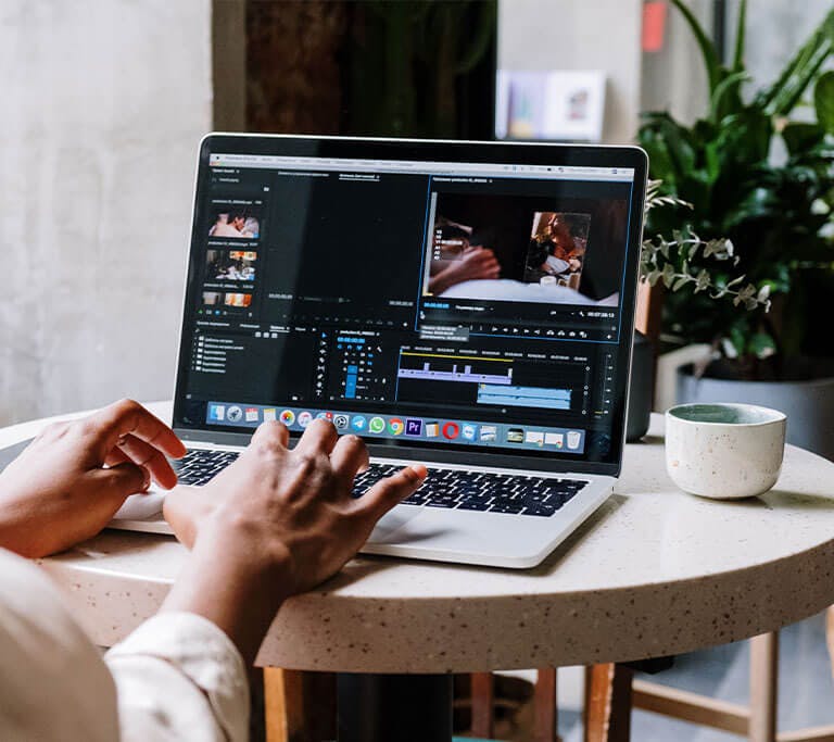 Over-the-shoulder view of a person using a MacBook laptop to edit video on a small, round table with plants.