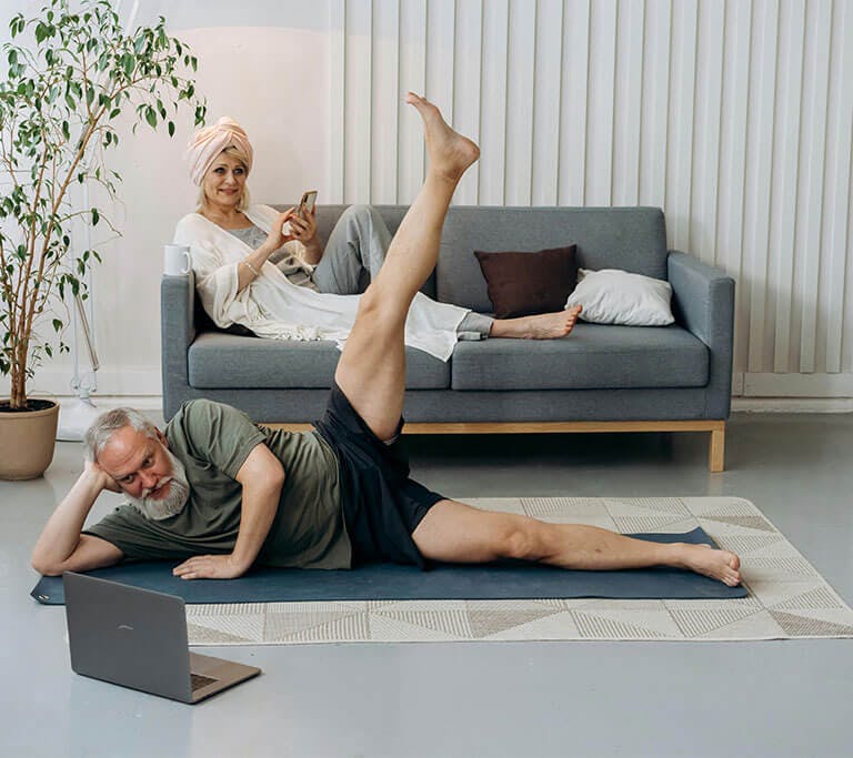 A man with a white beard lies on a blue mat on the floor and watches his laptop screen while a smiling woman lounges on the couch behind him, using her phone.