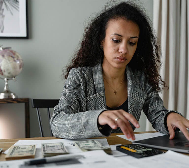 A person works on a small business budget at a desk, comparing paperwork while using a laptop and calculator.
