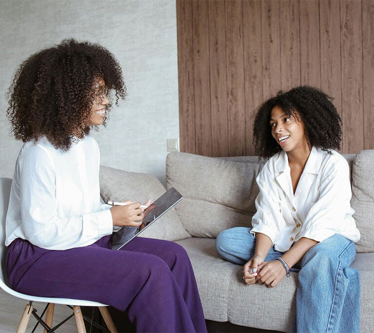 Two people with curly hair sit and talk in a cozy, modern living room. One person is seated on a white chair holding a tablet and pen, while the other sits on a beige couch smiling and engaging in conversation.
