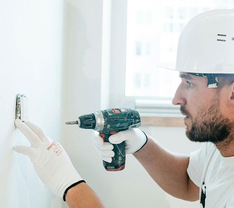 A person wearing a white shirt, gloves and a safety helmet drills a hole in a wall using a power drill.