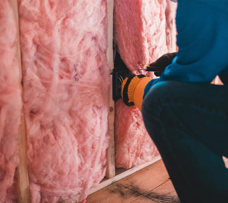 A contractor wearing jeans, a blue shirt and safety gloves squats and adjusts exposed pink wall insulation.