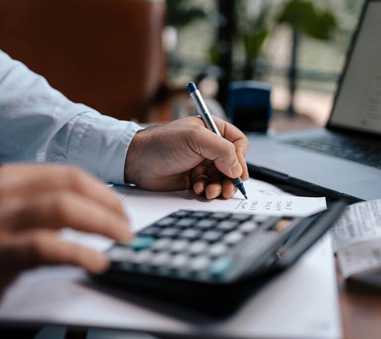 person uses a calculator while writing down expenses in a room with lots of windows and a large houseplant.