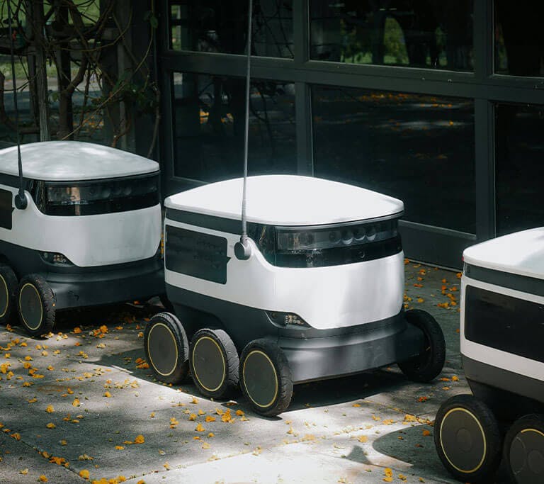 Three white package-delivery robots roll down the street, one after the other in a line.