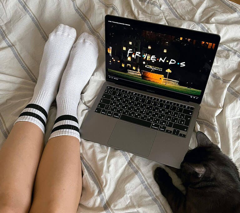 A person wearing white sits on a bed next to a black cat while watching Friends on their laptop.