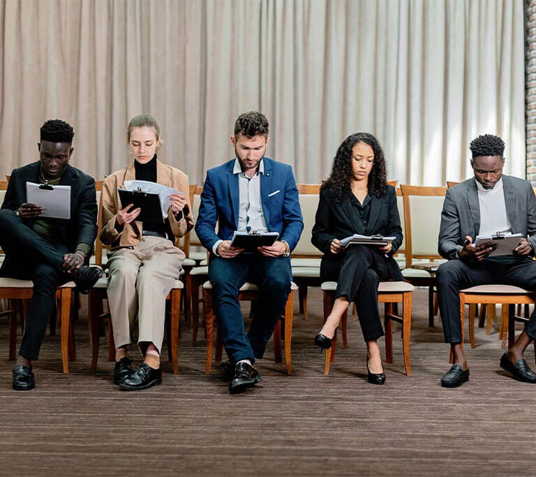 Five professionally dressed people sit in a row of chairs, reviewing documents on clipboards in what appears to be a waiting room or conference setting.