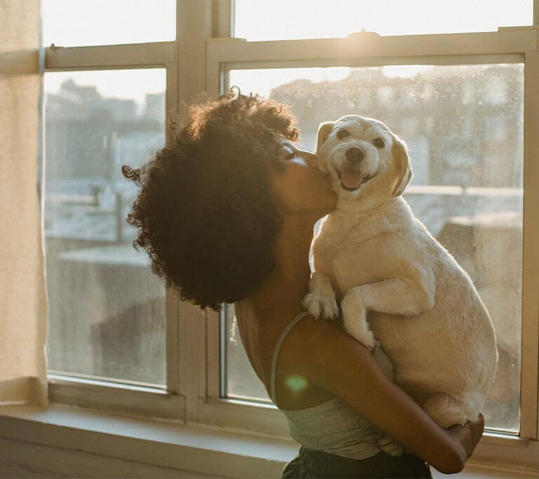 A woman is kissing her dog inside her home in front of a window.