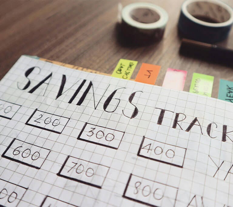 Graph paper with “Savings Trackers” and “Yay!” handwritten on it, along with several numbers and boxes, sits on a wooden table next to tape and marker.