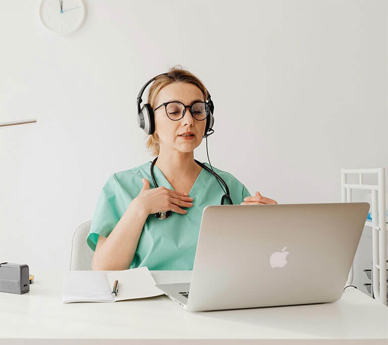 A physician is wearing a headset and sitting in a room on a video call on a Macbook computer.