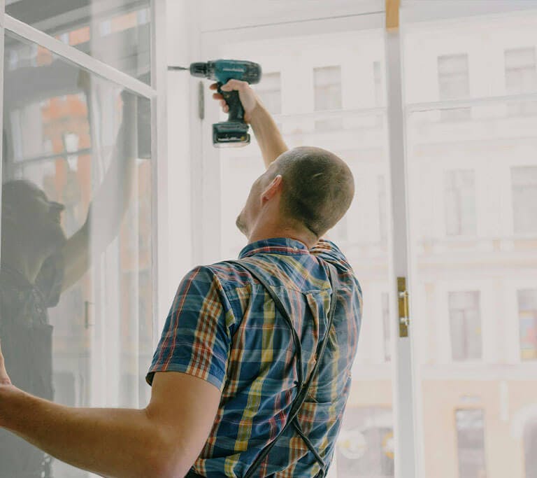 A “handyman” wearing a blue checkered shirt presses a drill against a window frame.