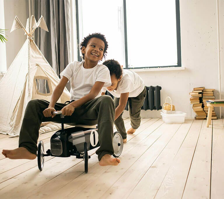 A child pushes a grinning child who is riding around on a car toy inside a beautiful playroom with a stack of books, a majesty palm houseplant and a miniature teepee.