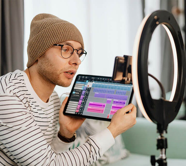 An influencer in a striped long-sleeve shirt wearing a taupe beanie and rounded glasses sits on a mint-green couch and holds up a tablet in front of a circle light with a mobile phone recording their demonstration.