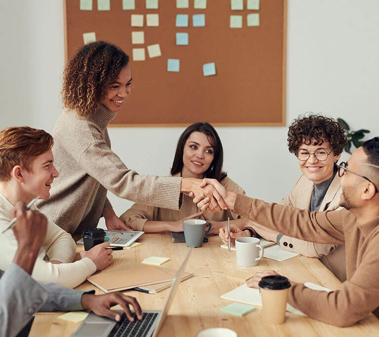 A group of six smiling professional young adults sit at a wooden table and shake hands during a meeting.