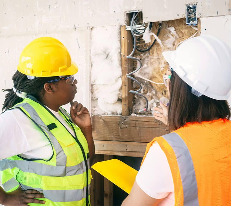 Two people wearing high-visibility safety vests and hard hats analyze an area of a wall with exposed insulation.