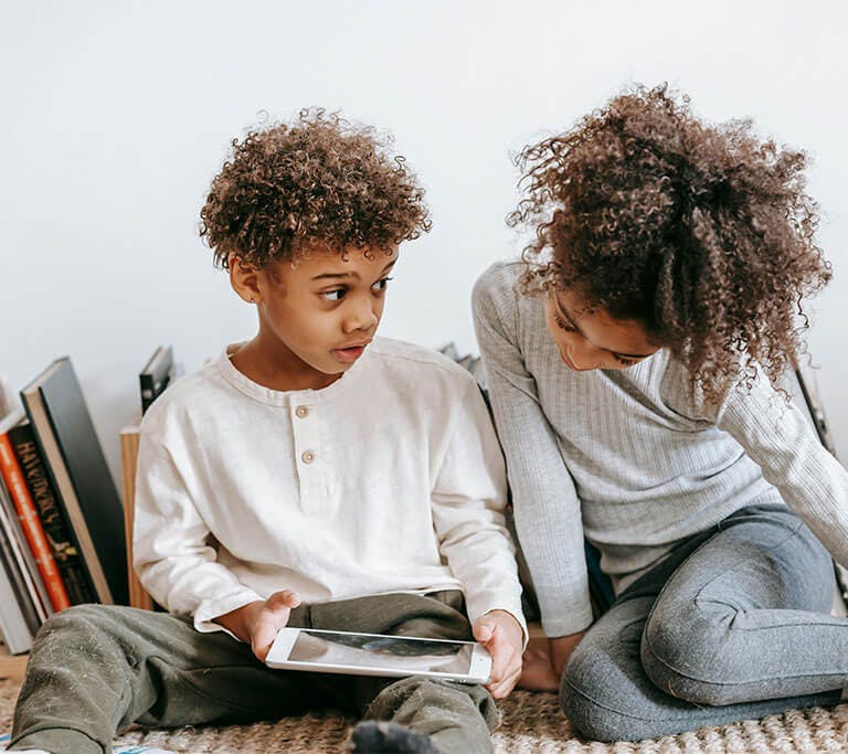 Two kids sit in front of their books on the carpet while using a tablet.