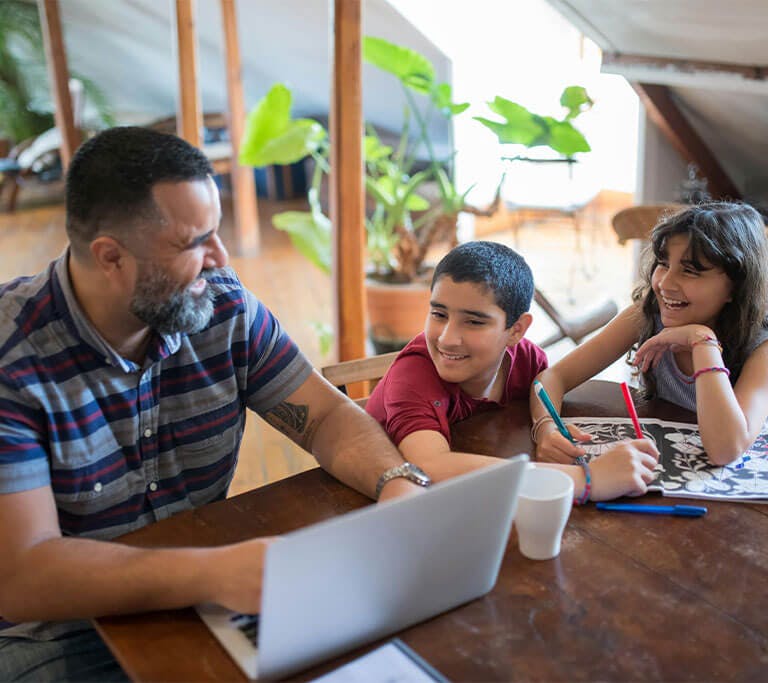 A father and two children at a table smile while using a laptop together.