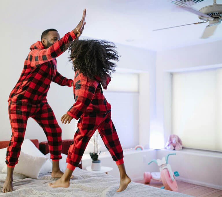 A father and daughter wearing matching red-and-black checkered pajamas high-five while jumping on a bed in a bedroom with a ceiling fan and pink tricycle.
