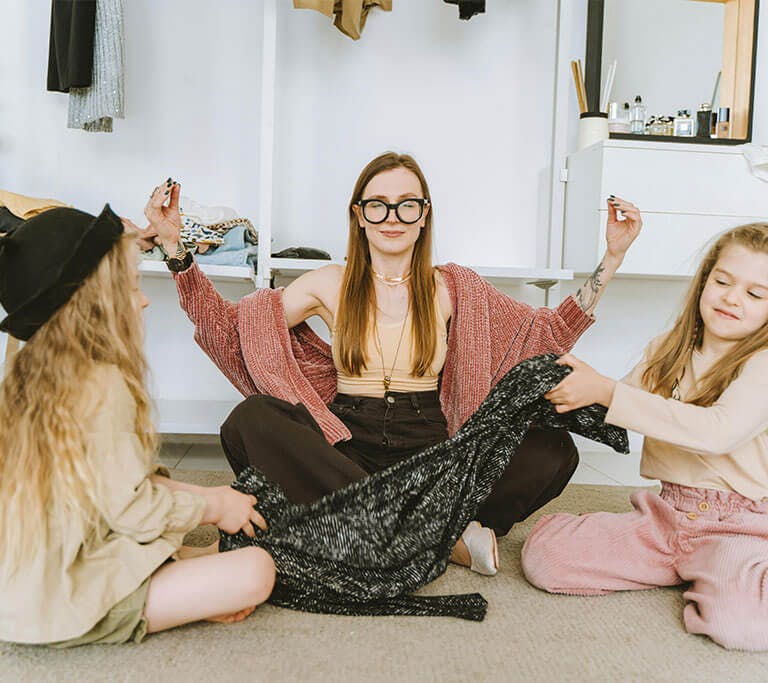 A mother tries to happily meditate while her two children tug on a blanket on the floor.